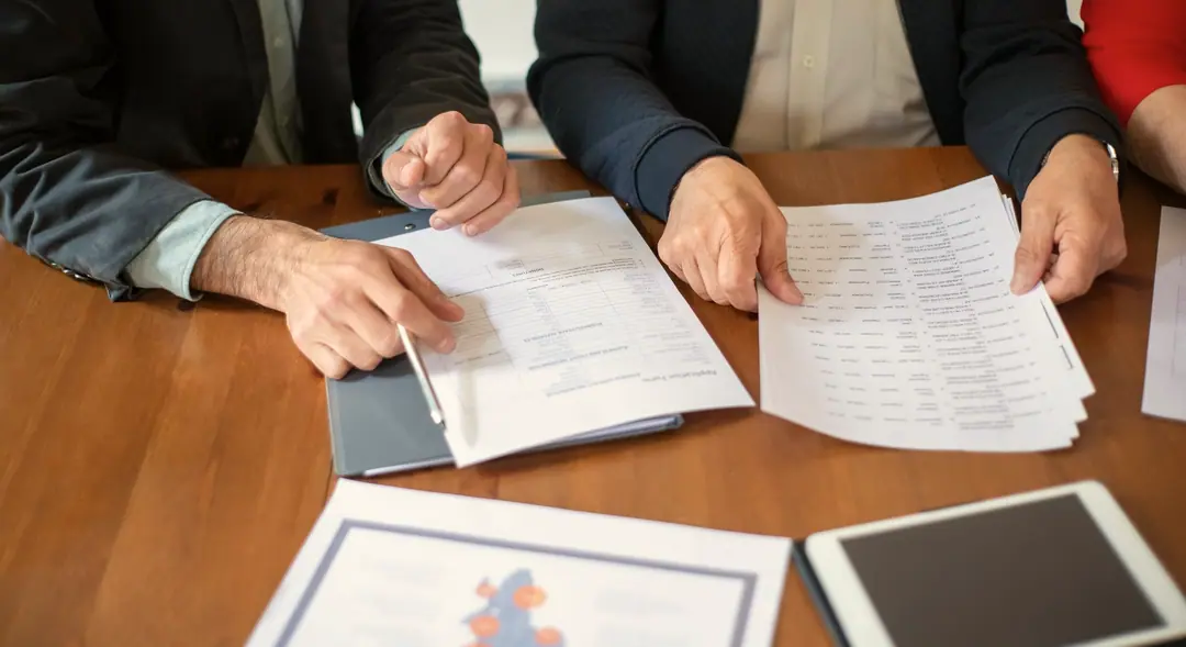 Двама души преглеждат печатни таблици и документи на дървена маса